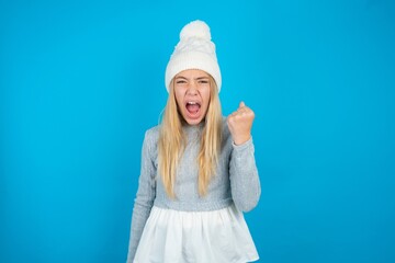 Teen caucasian girl wearing blue knitted sweater and woolly hat angry and mad raising fist...