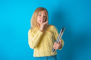 Beautiful kid girl wearing yellow sweater holding notebook makes silence gesture, keeps finger over lips. Silence and secret concept.