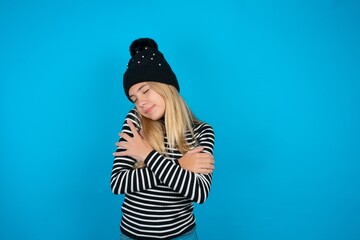 Teen caucasian girl wearing striped sweater and woolly hat Hugging oneself happy and positive,...