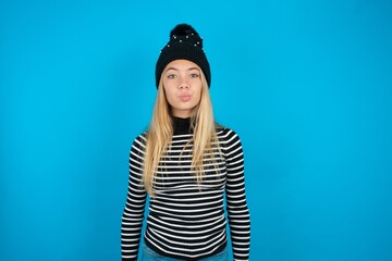 Shot of pleasant looking Teen caucasian girl wearing striped sweater and woolly hat , pouts lips,...