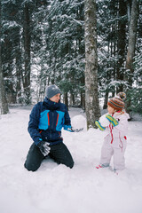 Dad and little girl show each other big snowballs for a snowman in a snowy forest