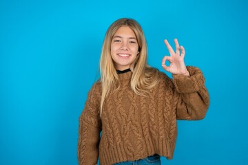 beautiful caucasian teen girl wearing yellow sweater hold hand arm okey symbol toothy approve...