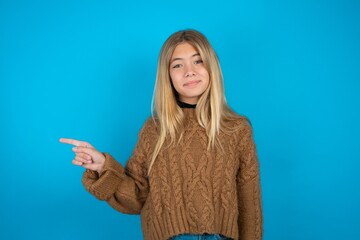 Fototapeta premium Positive beautiful caucasian teen girl wearing brown sweater with satisfied expression indicates at upper right corner shows good offer suggests to click on link