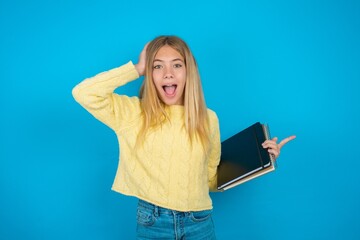 Surprised beautiful caucasian teen girl wearing yellow sweater pointing at empty space holding hand on head