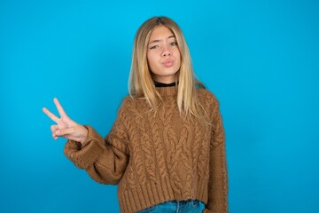 beautiful caucasian teen girl wearing brown sweater makes peace gesture keeps lips folded shows v...