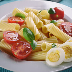 Grooved pasta with boiled egg, tomatoes and basil on a white plate. Square.