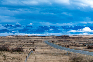 Photograph of a road heading towards a mountain range through a rural area with low level grey clouds on the South Island of New Zealand