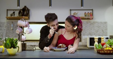 Indian Asian Hindu happy young couple hold spoon eat tasty sweet cake pastry snacks food sitting kitchen enjoy indoor house party adult cute boy girl male female lady talk gossip make fun joy day home