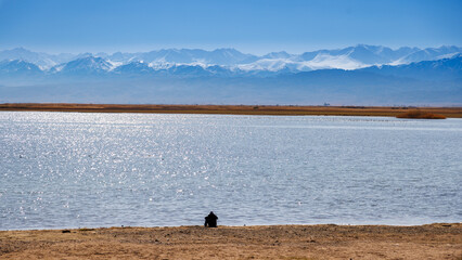 alone human silhouette sitting on the beach of blue calm water in Issyk-Kul lake with mountains on background at autumn afternoon. - Powered by Adobe