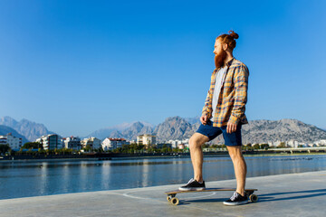 handsome redhaired man with long beard skateboarding near river at sunny summer day