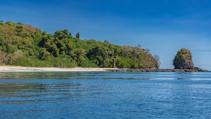 The tropical island is covered with green vegetation. A picturesque rock is nearby. A white sandy beach, ripples and highlights on the blue water. The boat is moored near the shore. Azure sky. 