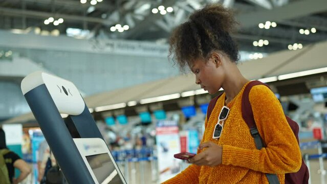 African woman holding passport using self Check-in kiosk machine getting airline ticket boarding pass at airport terminal. People travel on holiday vacation and global airplane transportation concept.