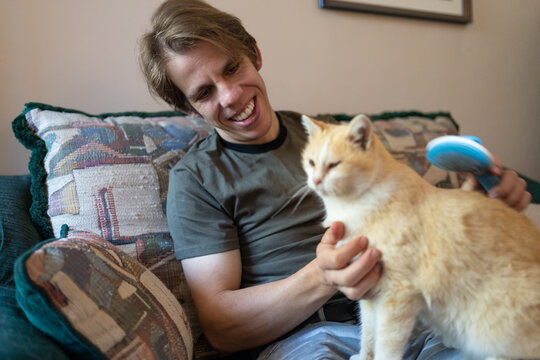 In a cozy living room at home, a young adult man carefully brushes a cute kitten, displaying affectionate grooming.