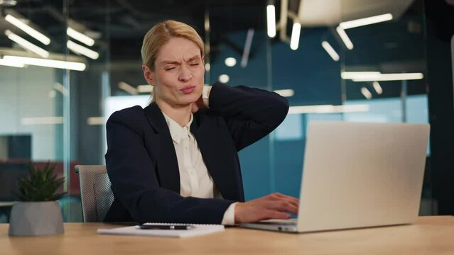 Exhausted businesswoman seated at her desk, enduring intense neck pain and alleviating muscle tension through gentle rubbing. Entrepreneur struggles with spinal tension in office, sedentary lifestyle.