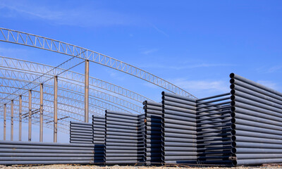 Pile of many metal roof beams for installing on top of large industrial factory building outline structure in construction site area against blue sky background