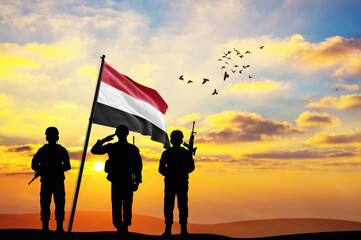 Silhouettes of soldiers with the Yemen flag stand against the background of a sunset or sunrise....