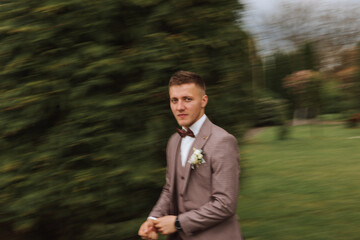 The groom in a brown suit adjusts his jacket, poses against the background of green trees. Wedding portrait.