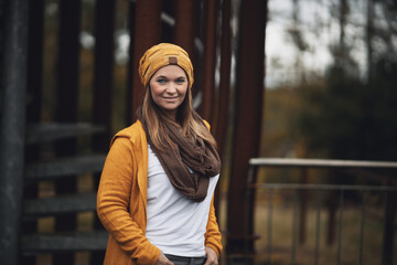 Woman brunette in knitted wool with hat and brown scarf, standing in front of a steel frame in the autumn forest.