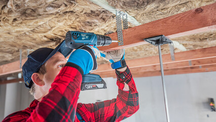 Lowering and insulate the ceiling. Fixing a wooden prism using suspend tin sheet to the load-bearing wooden structure of the ceiling. The worker works with a cordless drill. - obrazy, fototapety, plakaty