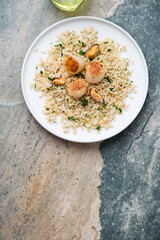 White plate with scallops, brown rice and mussels, flat lay on a beige and grey granite background, vertical shot, copy space