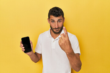 Young Hispanic man shows phone screen pointing with finger at you as if inviting come closer.
