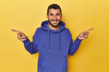 Young Hispanic man on yellow background pointing to different copy spaces, choosing one of them,...