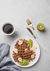 Belgian vegetarian waffles with kiwi, blueberries and banana slices on a white plate with a cup of coffee on a light background.
