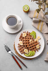 Belgian chocolate waffles with kiwi, blueberries and banana slices on a white plate with a cup of coffee on a light background.
