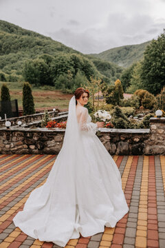 A red-haired bride in an elegant dress with long sleeves and a beautiful ornament in her hair, poses with her shoulders turned to the camera on the background of mountains. Long train of the dress.