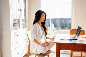 Young cute Asian woman in white clothes is sitting on chair in lotus position and meditating....