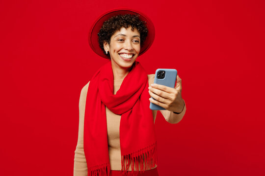 Young fun woman of African American ethnicity she wears beige sweater hat scarf doing selfie shot on mobile cell phone post photo on social network isolated on plain red background. Lifestyle concept.