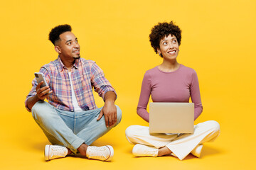 Full body young couple two friends family IT man woman wear purple casual clothes together sitting hold mobile cell phone use work on laptop pc computer look aside isolated on plain yellow background. - obrazy, fototapety, plakaty