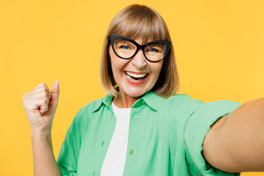 Close up elderly blonde woman 50s years old wear green shirt glasses casual clothes doing selfie shot pov on mobile cell phone do winner gesture isolated on plain yellow background. Lifestyle concept.