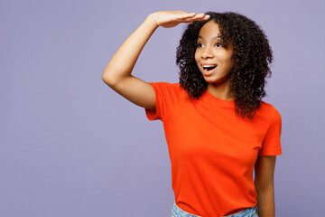 Little kid teen girl of African American ethnicity she wear orange t-shirt hold hand at forehead look far away distance isolated on plain pastel light purple background. Childhood lifestyle concept.