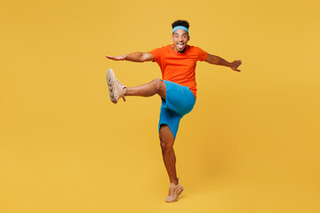 Fototapeta na wymiar Full body young fitness trainer sporty man sportsman wear orange t-shirt stand with outstretched hands raise up leg train in home gym isolated on plain yellow background Workout sport fit abs concept