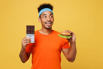 Young fitness trainer sporty man sportsman wear orange t-shirt hold fastfood burger chocolate bar look aside spend time in home gym isolated on plain yellow background. Workout sport fit abs concept. - obrazy, fototapety, plakaty