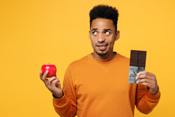 Young minded man wear sweatshirt casual clothes hold eat bar of chocolate, apple, look aside on area isolated on plain yellow background. Proper nutrition healthy fast food unhealthy choice concept. - obrazy, fototapety, plakaty