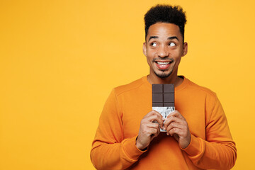 Young man he wear orange sweatshirt casual clothes hold eat bar of chocolate look aside on area isolated on plain yellow background studio. Proper nutrition healthy fast food unhealthy choice concept. - obrazy, fototapety, plakaty