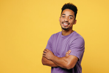 Side view young smiling happy man of African American ethnicity he wears purple t-shirt casual clothes hold hands crossed folded isolated on plain yellow background studio portrait. Lifestyle concept. - obrazy, fototapety, plakaty