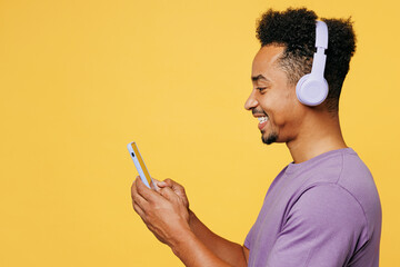 Side view young man of African American ethnicity wears purple t-shirt casual clothes use mobile cell phone listen to music in headphones isolated on plain yellow background studio. Lifestyle concept.