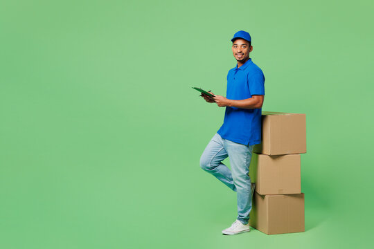 Full body side view delivery guy employee man wear blue cap t-shirt uniform workwear work as dealer courier near boxes hold clipboard paper document isolated on plain green background Service concept.