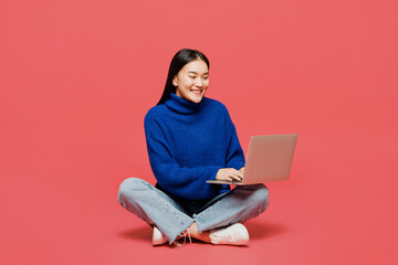 Full body young IT woman of Asian ethnicity wear blue sweater casual clothes sitting hold use work on laptop pc computer isolated on plain pastel light background studio portrait. Lifestyle concept.