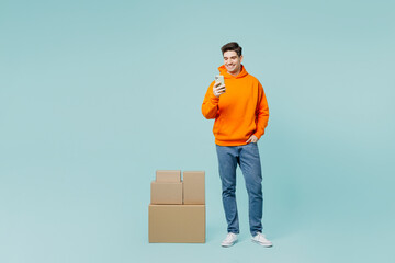 Full body young happy smiling man he wear orange hoody casual clothes stand near stack cardboard...