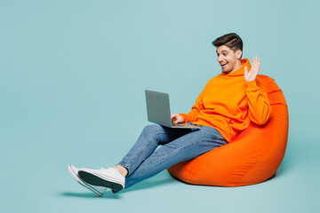 Full body young IT man wears orange hoody casual clothes sit in bag chair hold use work on laptop pc computer waving hand isolated on plain pastel light blue cyan color background. Lifestyle concept.