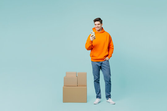 Full body young happy smiling man he wear orange hoody casual clothes stand near stack cardboard blank boxes holding use mobile cell phone isolated on plain blue color background. Lifestyle concept.