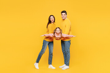 Full body young cool happy parents mom dad with child kid girl 7-8 years old wear pink sweater casual clothes hold daughter pov she is flying isolated on plain yellow background. Family day concept.