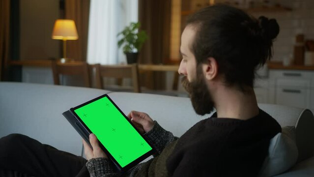 Bearded Hair Bun Man Scrolling And Tapping Center On Tablet With Green Screen Mock Up Display. Male Lyin On Sofa, Relaxing At Home. Close-up Over The Shoulder Shot