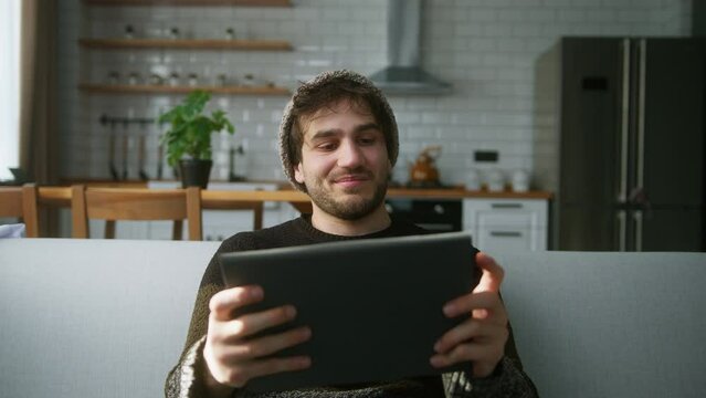 Young man with beanie sitting on sofa at home with kitchen background, playing excited concentrated engrossed in interesting mobil game with tablet computer