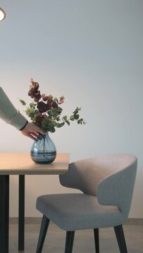 A man's hand places a glass blue vase on a wooden table. A series of upholstered chairs
