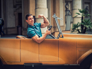 A young man is sitting in a fake vintage car, doing a bodybuilding bicep pose and smiling to the...